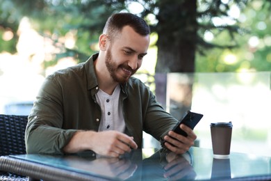 Photo of Happy man with paper cup using smartphone at outdoor cafe