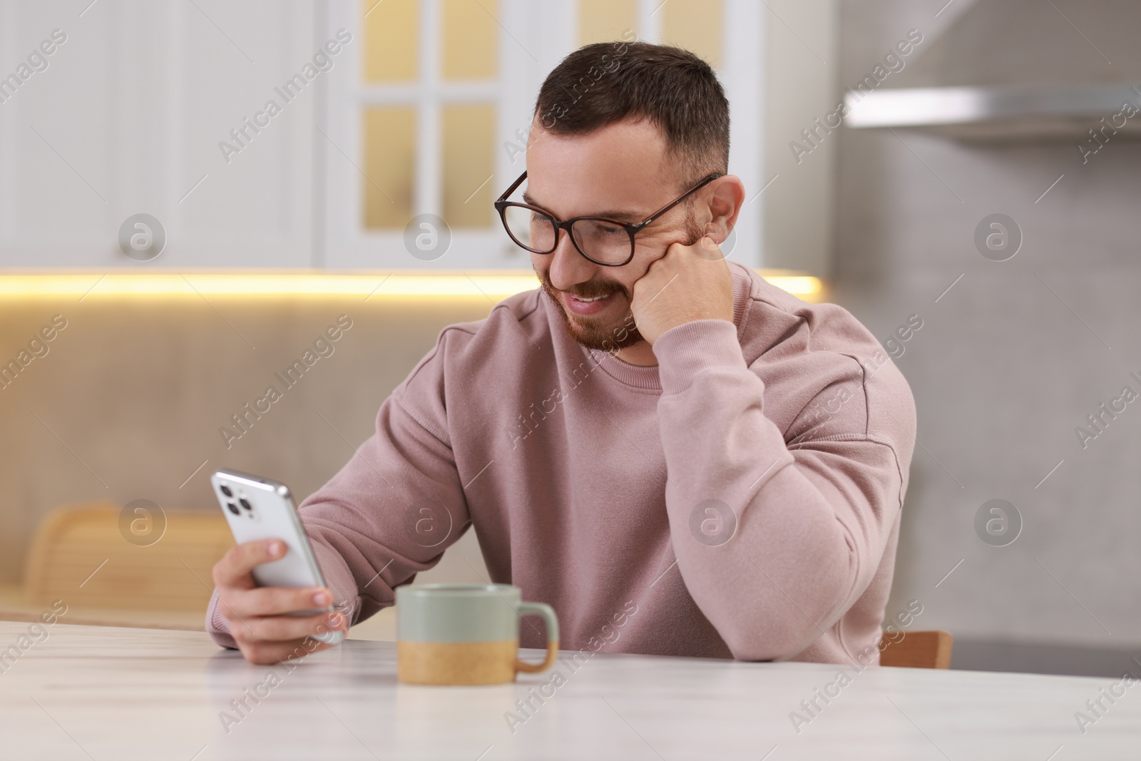 Photo of Happy man using smartphone at white table in kitchen