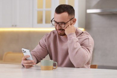 Happy man using smartphone at white table in kitchen