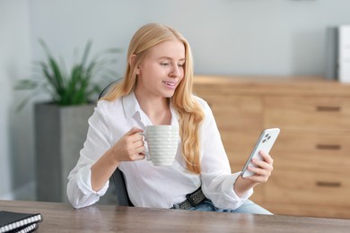 Photo of Smiling woman using smartphone and drinking coffee at table in office