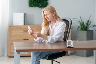 Photo of Smiling woman using smartphone and drinking coffee at table in office