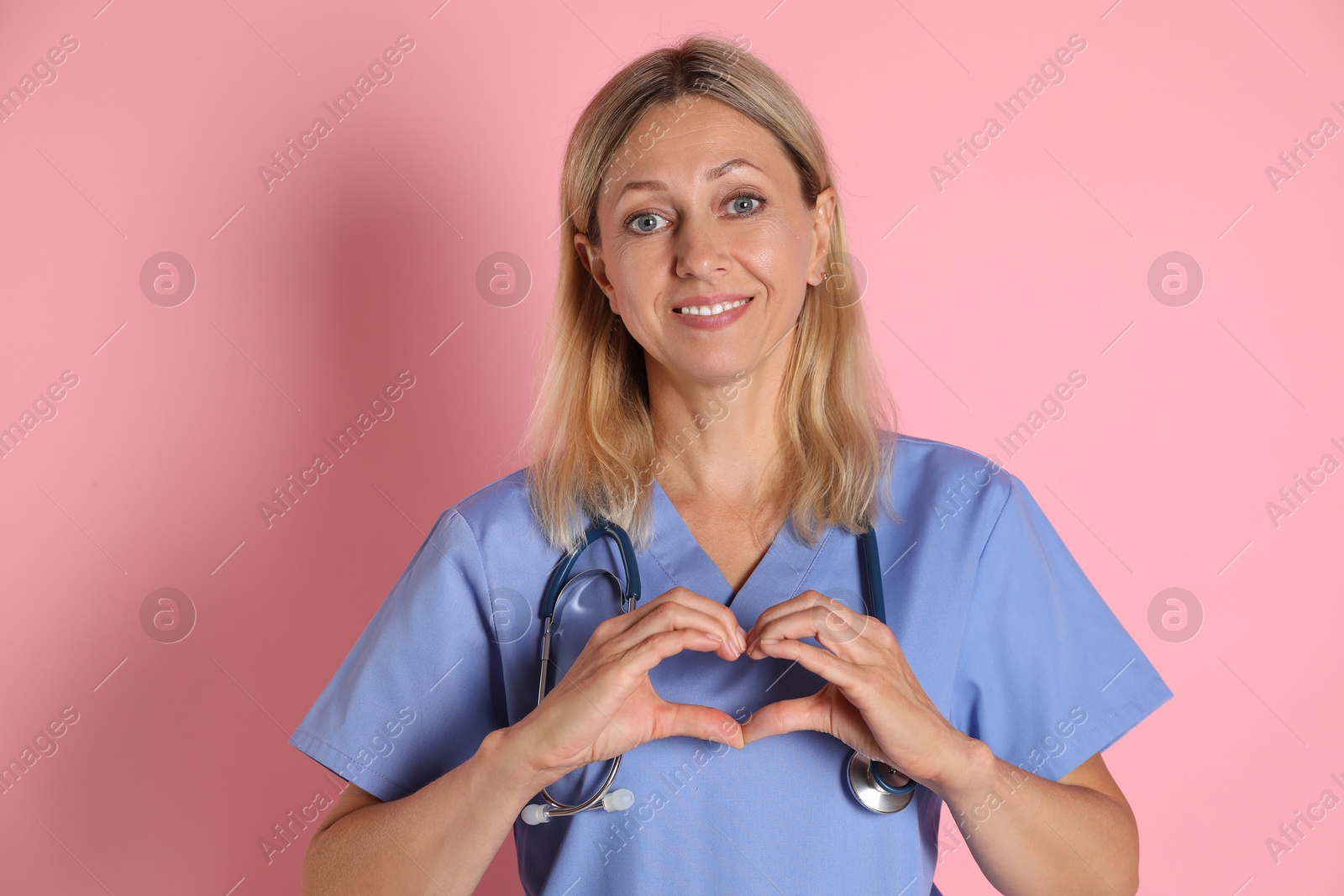 Photo of Doctor in medical uniform with stethoscope making heart with hands on pink background