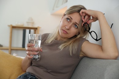 Photo of Menopause. Woman with glass of water suffering from headache on sofa at home