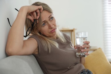 Photo of Menopause. Woman with glass of water suffering from headache on sofa at home