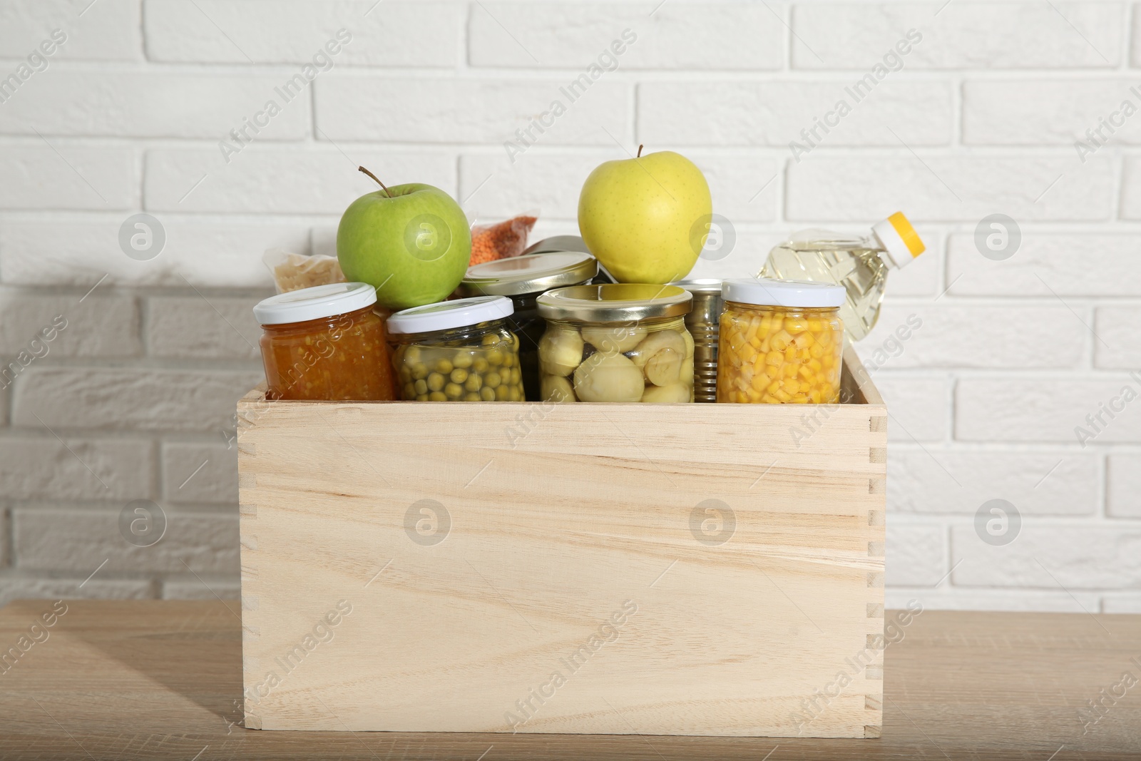 Photo of Different food products for donation in wooden crate on table near white brick wall