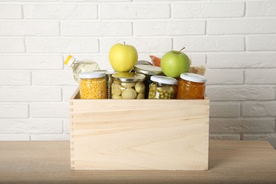 Photo of Different food products for donation in wooden crate on table near white brick wall