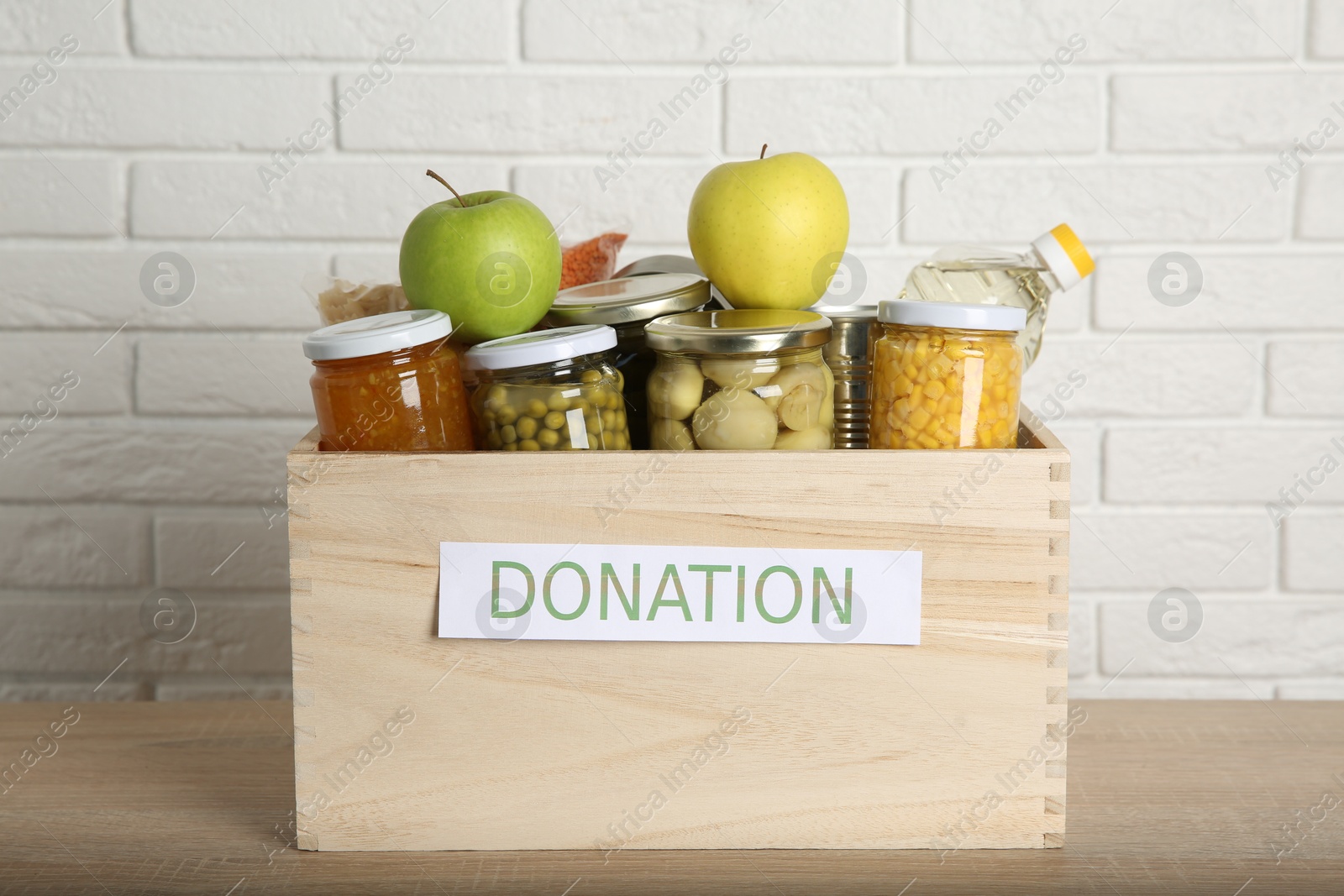 Photo of Different food products for donation in wooden crate on table near white brick wall