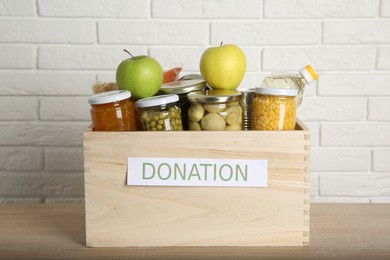 Photo of Different food products for donation in wooden crate on table near white brick wall