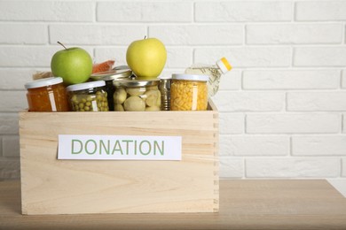 Photo of Different food products for donation in wooden crate on table near white brick wall