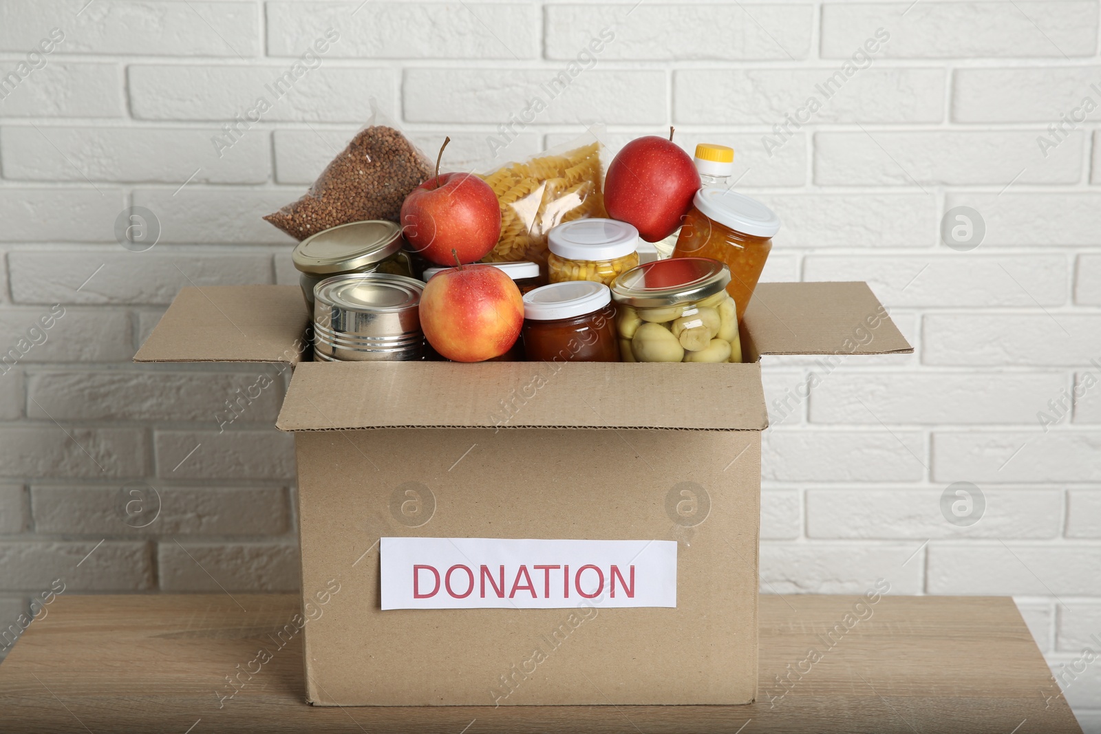 Photo of Different food products for donation in box on wooden table near white brick wall