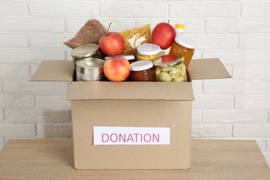 Photo of Different food products for donation in box on wooden table near white brick wall