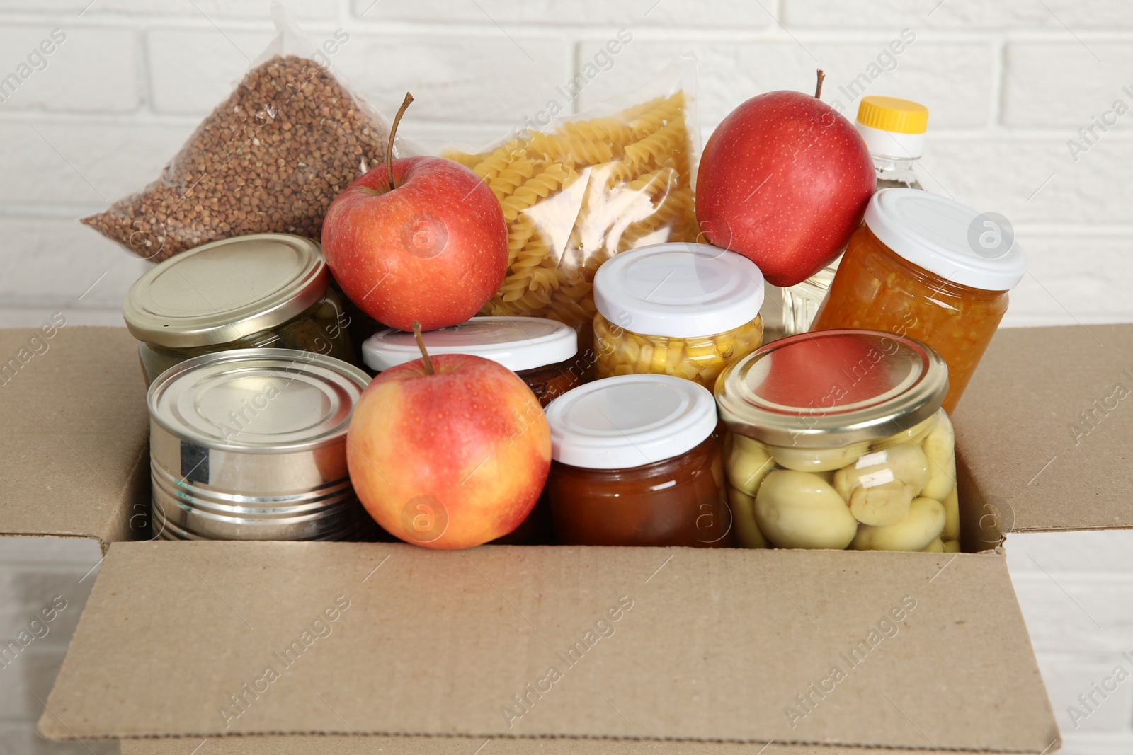 Photo of Different food products for donation in box near white brick wall, closeup