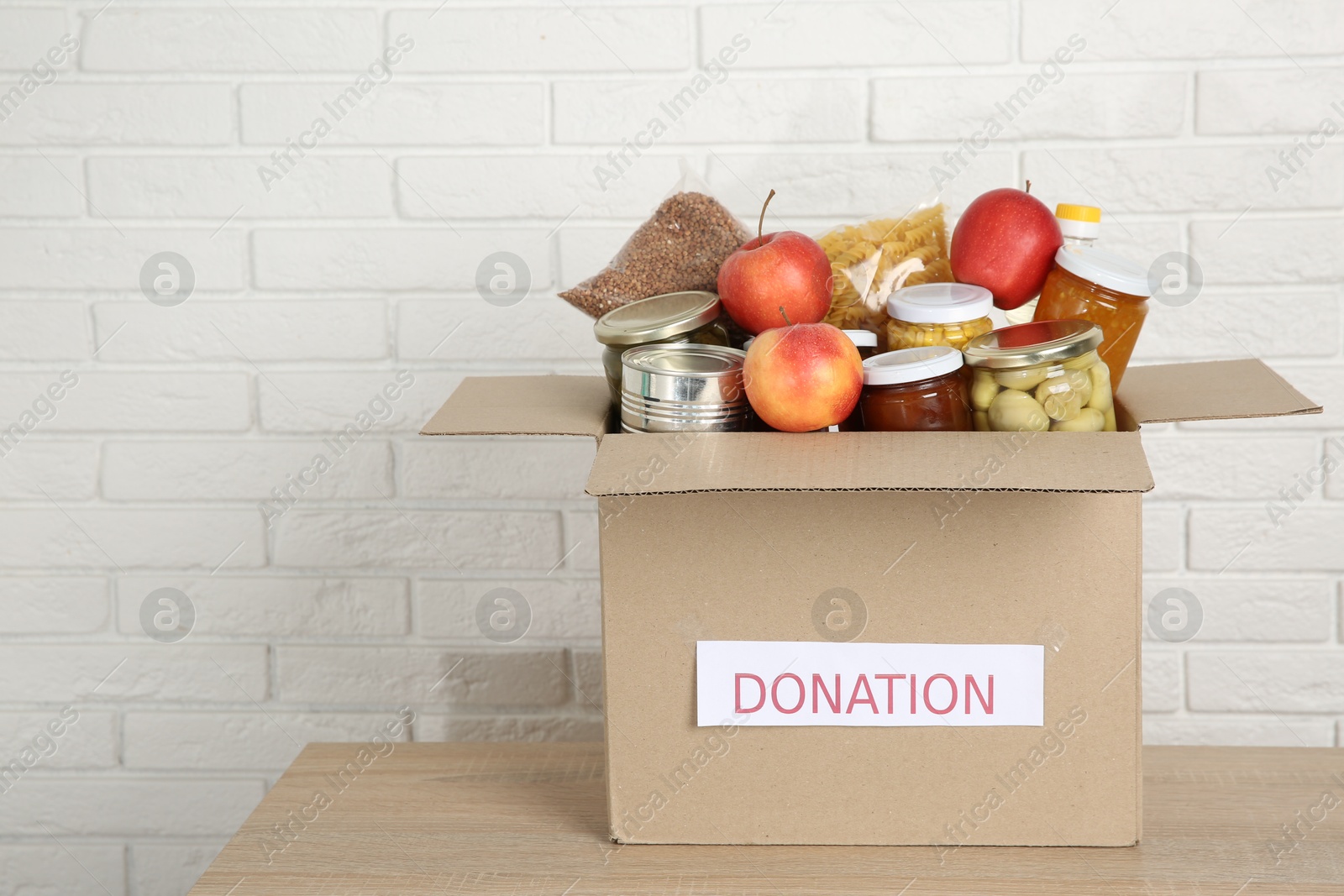 Photo of Different food products for donation in box on wooden table near white brick wall