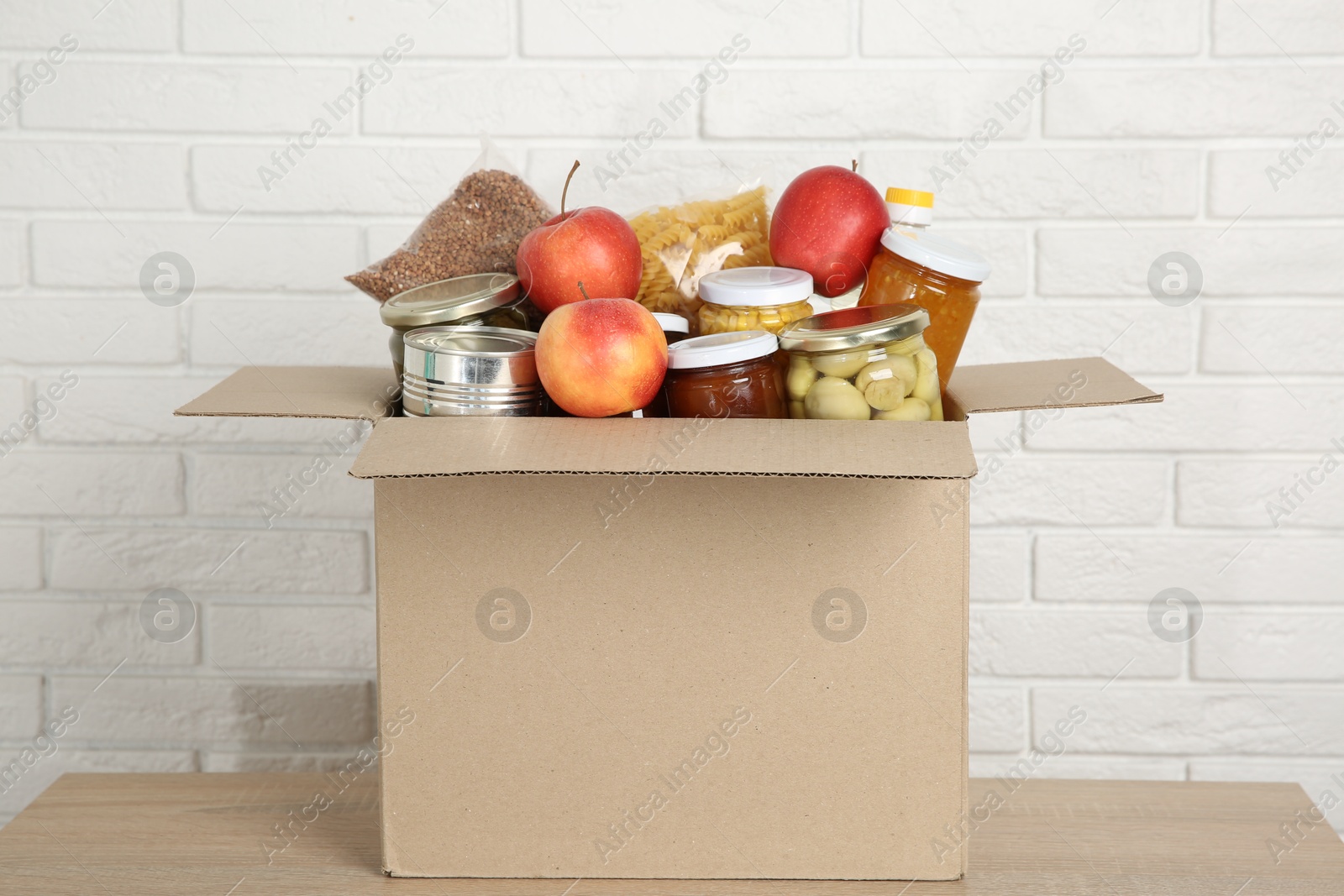 Photo of Different food products for donation in box on wooden table near white brick wall