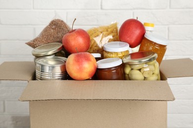 Photo of Different food products for donation in box near white brick wall, closeup