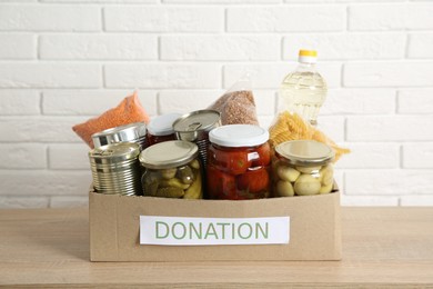 Photo of Different food products for donation in box on wooden table near white brick wall