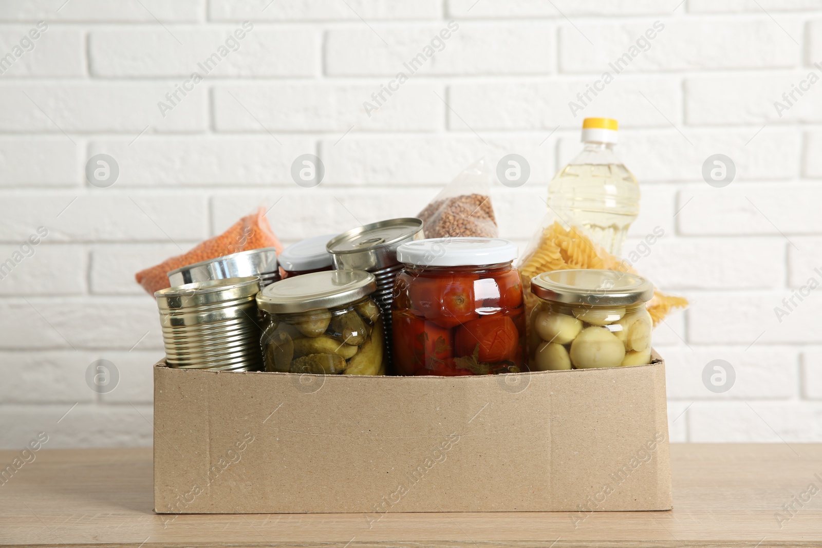 Photo of Different food products for donation in box on wooden table near white brick wall