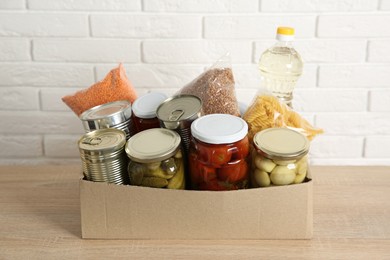 Photo of Different food products for donation in box on wooden table near white brick wall