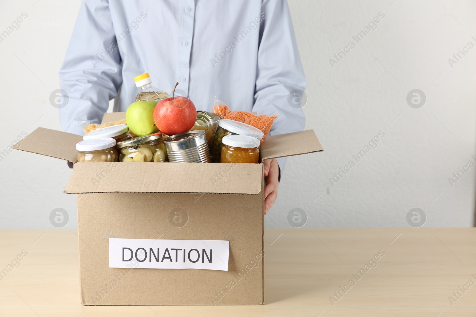 Photo of Food donation. Woman with box of different products at wooden table indoors, closeup