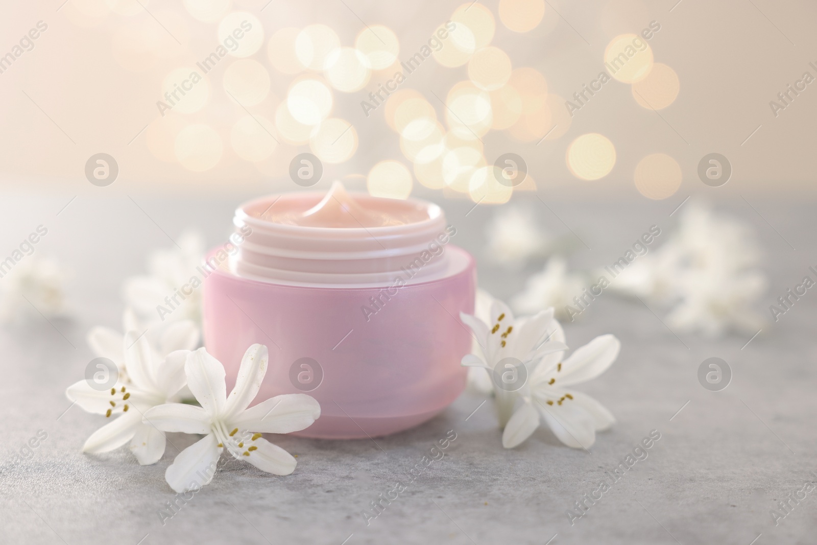 Photo of Cream in jar and jasmine flowers on grey table against light background with blurred lights, closeup