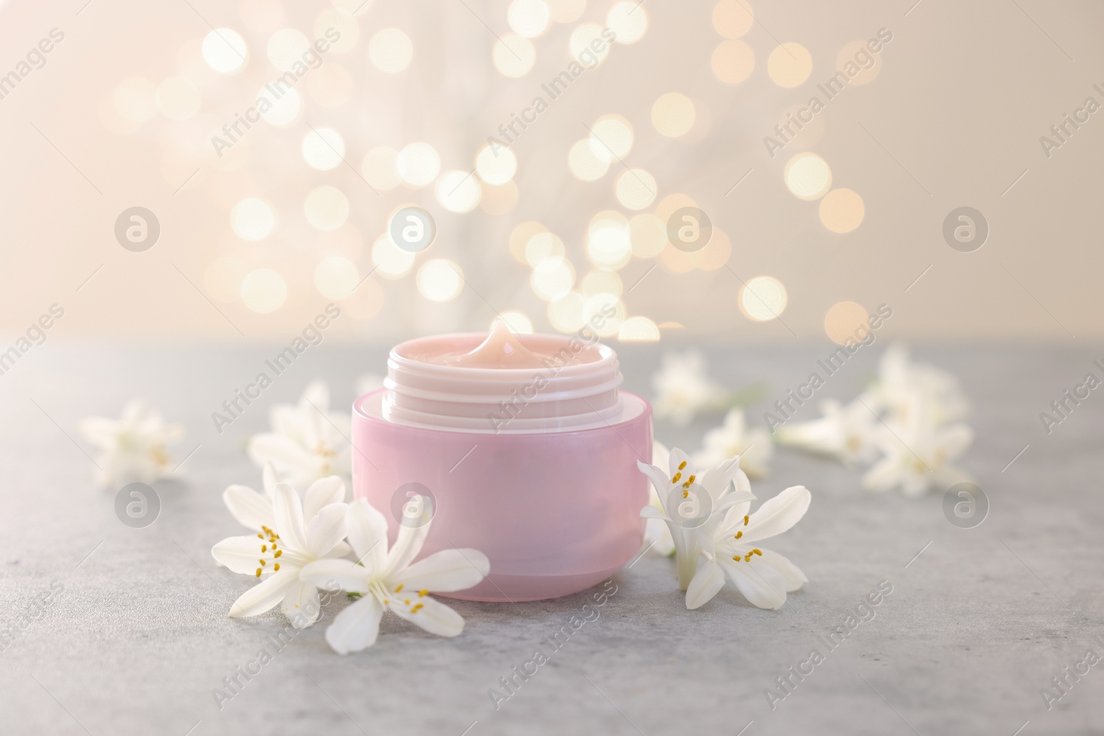 Photo of Cream in jar and jasmine flowers on grey table against beige background with blurred lights, closeup