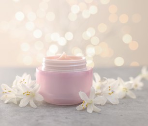 Photo of Cream in jar and beautiful jasmine flowers on grey table against beige background with blurred lights, closeup