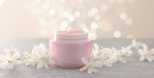 Photo of Cream in jar and beautiful jasmine flowers on grey table against beige background with blurred lights, closeup