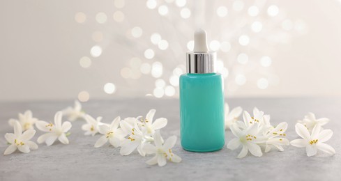 Photo of Cosmetic product in bottle and beautiful jasmine flowers on grey table against beige background with blurred lights