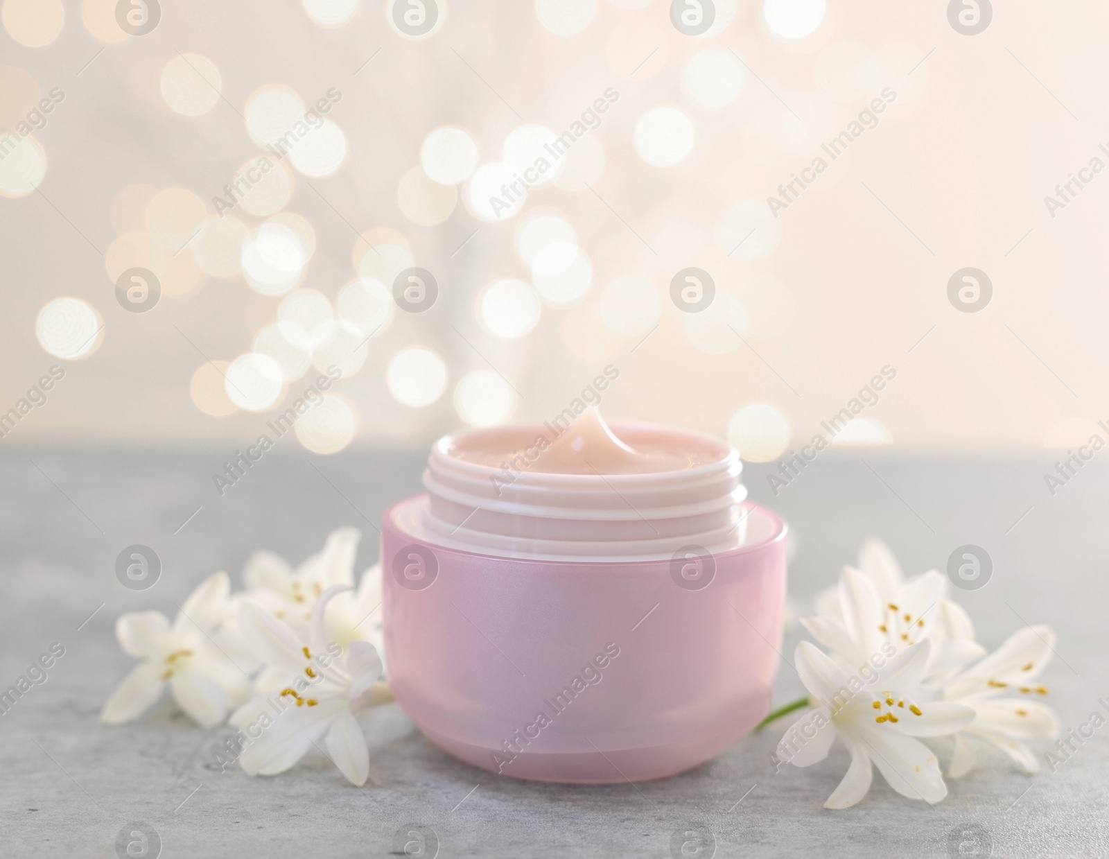 Photo of Cream in jar and beautiful jasmine flowers on grey table against light background with blurred lights, closeup. Space for text