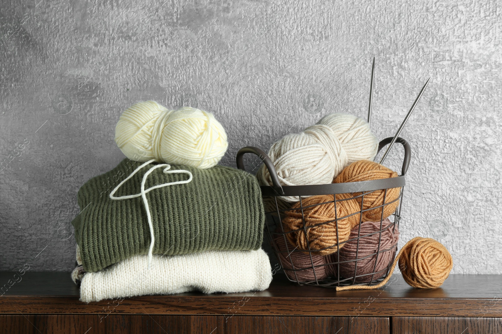 Photo of Basket with colorful yarns, knitting needles and stack of sweaters on wooden shelf indoors