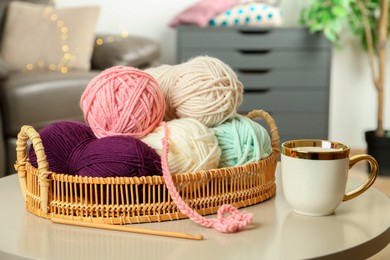 Wicker basket with colorful yarns, crochet pattern, hook and cup on coffee table indoors