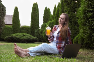 Woman with juice sitting near laptop on green lawn in park