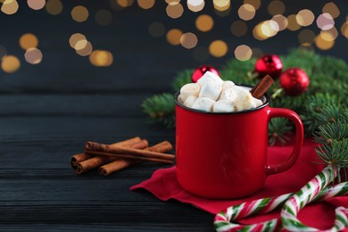 Photo of Tasty hot cocoa drink with marshmallows in mug, candy canes and Christmas decor on black wooden table, closeup. Space for text