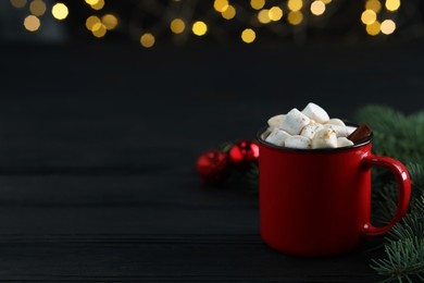 Photo of Tasty hot cocoa drink with marshmallows in mug and fir branches on black wooden table, closeup. Space for text