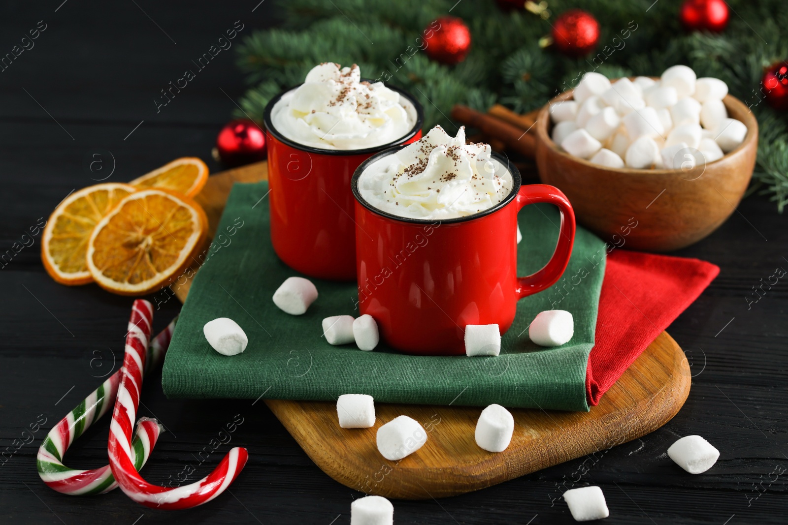 Photo of Tasty hot cocoa drinks with whipped cream in mugs, marshmallows and candy canes on black wooden table, closeup