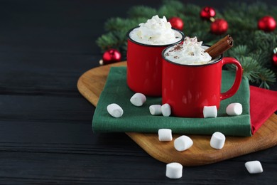 Photo of Tasty hot cocoa drinks with whipped cream in mugs and marshmallows on black wooden table, closeup. Space for text