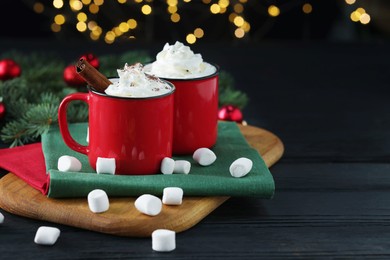 Photo of Tasty hot cocoa drinks with whipped cream in mugs and marshmallows on black wooden table, closeup. Space for text