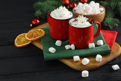 Photo of Tasty hot cocoa drinks with whipped cream in mugs and marshmallows on black wooden table, closeup