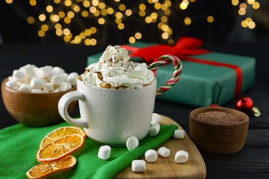 Photo of Tasty hot cocoa drink with whipped cream in cup and marshmallows on black wooden table, closeup