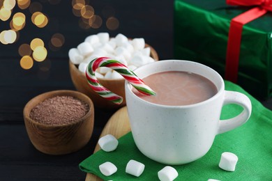 Photo of Tasty hot cocoa drink with candy cane in cup and marshmallows on black wooden table, closeup