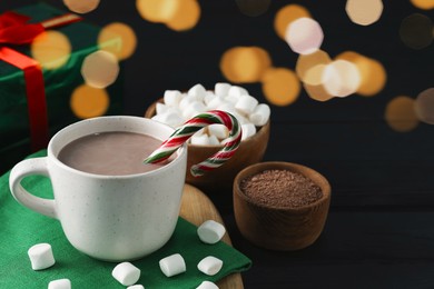 Photo of Tasty hot cocoa drink with candy cane in cup and marshmallows on black wooden table, closeup