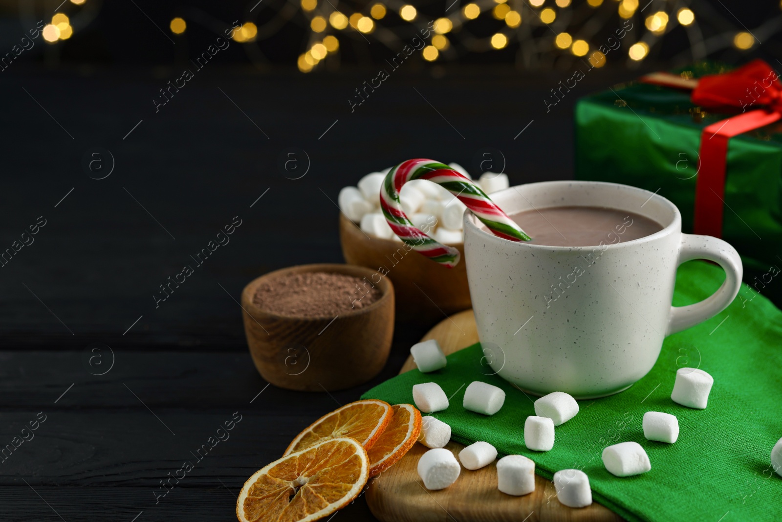 Photo of Tasty hot cocoa drink with candy cane in cup, dry orange slices and marshmallows on black wooden table, closeup. Space for text