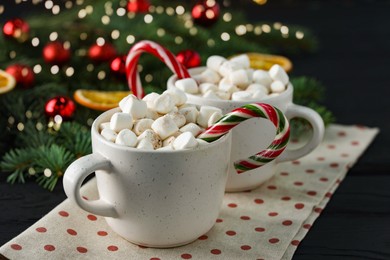 Photo of Tasty hot cocoa drinks with candy canes and marshmallows in cups on black wooden table, closeup