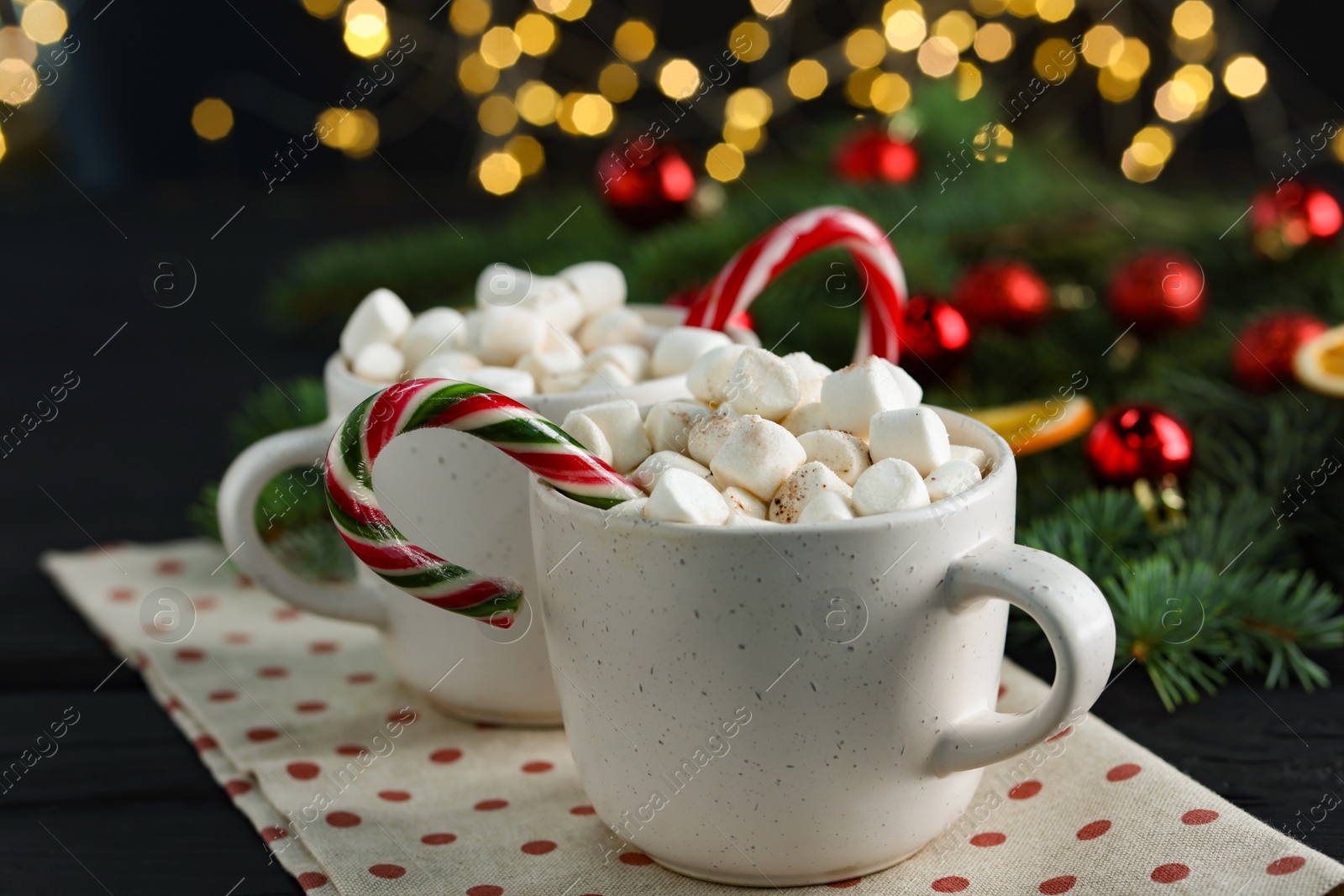 Photo of Tasty hot cocoa drinks with candy canes and marshmallows in cups on black wooden table, closeup