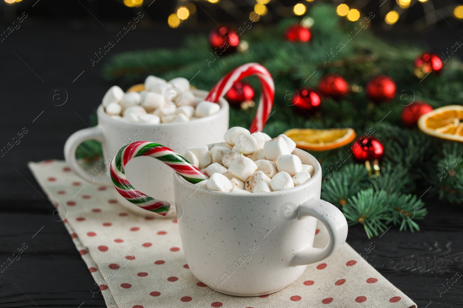 Photo of Tasty hot cocoa drinks with candy canes and marshmallows in cups on black wooden table, closeup