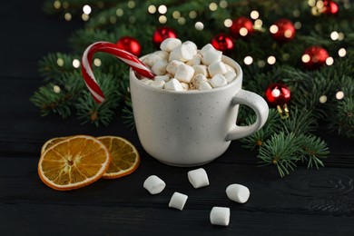 Photo of Tasty hot cocoa drink with candy cane, marshmallows in cup and dry orange slices on black wooden table, closeup