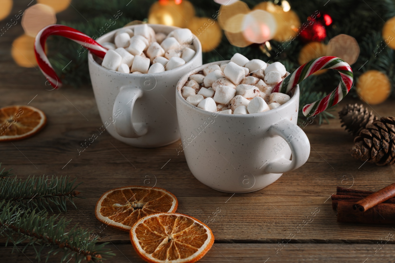 Photo of Tasty hot cocoa drinks with candy canes, marshmallows in cups and dry orange slices on wooden table, closeup