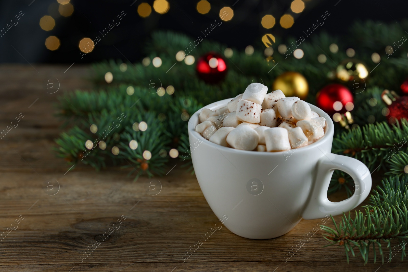 Photo of Tasty hot cocoa drink with marshmallows, cinnamon stick in cup and fir branches on wooden table, closeup. Space for text