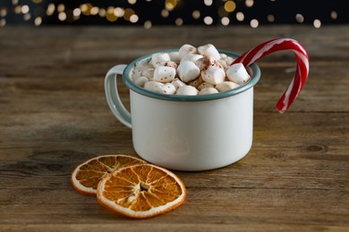 Photo of Tasty hot cocoa drink with marshmallows, candy cane in cup and dry orange slices on wooden table, closeup