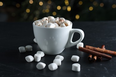 Photo of Tasty hot cocoa drink with marshmallows in cup and cinnamon sticks on black textured table